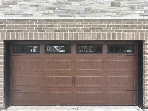 Brown garage door with rectangular panels and small windows at the top, set in a brick wall with stonework above.