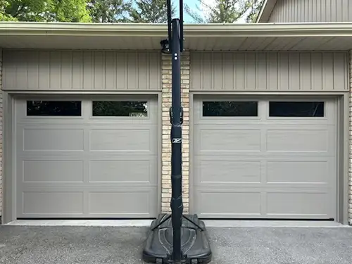 Two white garage doors on a brick house, separated by a portable basketball hoop.