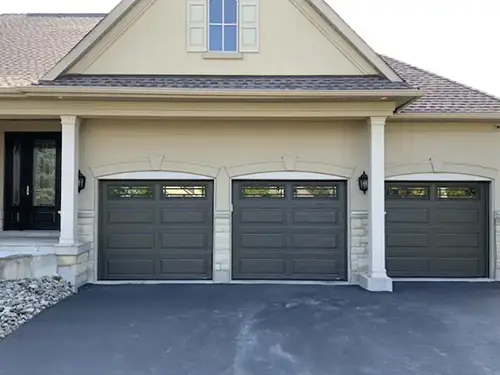 Three-car garage with dark grey doors and upper windows on a beige house. Two columns and stone accents frame the entrance.